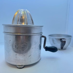 May include: Vintage metal citrus juicer with a silver finish. The juicer has a ribbed reamer, a handle with a black grip, and a separate small bowl. The juicer has an embossed emblem that reads "Home Service Bureau Canadian Home."