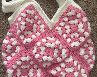 Puede incluir: Bolso tote de crochet hecho a mano con un patrón de cuadrados de la abuela. El bolso presenta una combinación de colores rosa y blanco, con una correa y ribete blancos. El bolso está diseñado con una forma redondeada y una abertura en forma de V.