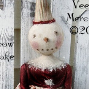 Pode incluir: Uma boneca de boneco de neve feita à mão vestindo um vestido de veludo vermelho com uma borda de renda branca e uma saia estampada em branco e vermelho que diz "Believe". A boneca está sentada em um banco de madeira branco com uma cerca branca de madeira no fundo. A boneca tem um chapéu cônico branco e um rosto pintado com bochechas rosadas.