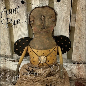 May include: A handmade rag doll dressed as a bee. The doll has a brown face with black eyes and a yellow and brown dress with black polka dots. The doll is sitting on a wooden surface with the text "Aunt Bee" written on the surface.