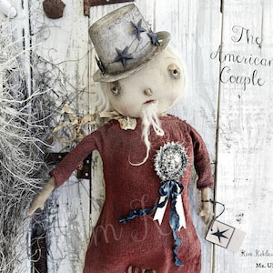May include: A handmade rag doll dressed in a red dress with a blue ribbon and a white hat with stars. The doll has a white beard and is holding a blue and white rosette. The doll is standing in front of a white wooden background. The text "The American Couple" is written in black above the doll. The text "Kim Koeller 2015 Mr. USA" is written in black below the doll.