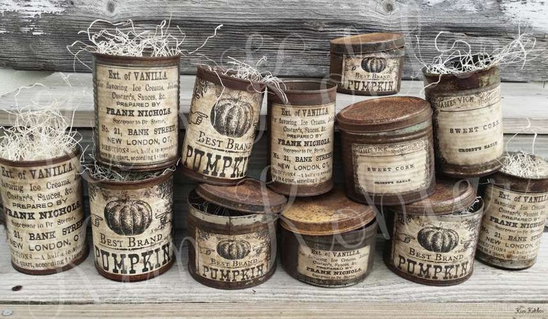 May include: A collection of vintage-style metal containers with aged labels. Some cans feature "Best Brand Pumpkin" and "Sweet Corn" labels. The containers are filled with decorative dried grass, creating a rustic, antique aesthetic.