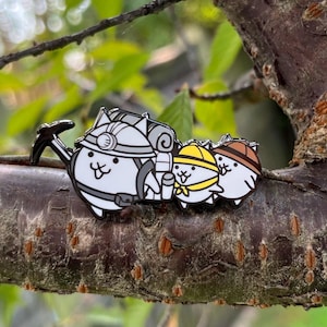 May include: A detailed enamel pin featuring three cartoon cat characters dressed as miners. The first cat wears a hard hat and carries a pickaxe and backpack. The other two cats wear yellow and brown hats. The pin is set against a blurred natural background.