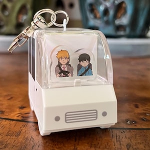 May include: White plastic keychain shaped like a van with a clear plastic window. The window shows two cartoon characters inside the van. The characters are wearing school uniforms and have black hair and brown hair.