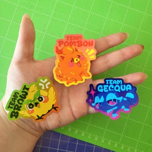May include: Three colorful stickers featuring cartoon characters. One is an orange dog with the text "TEAM POMBON". Another is a yellow bird with the text "TEAM BROWT". The third is a blue creature with the text "TEAM GECQUA".