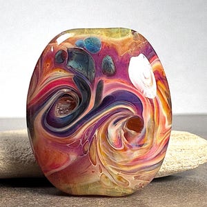 May include: A smooth, oval-shaped glass bead featuring a swirling, marbled pattern of vibrant colors including magenta, deep purple, orange, yellow, and hints of blue. The surface has a glossy finish with translucent areas and distinct color layers.