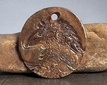 May include: A circular bronze-toned pendant featuring a detailed embossed relief of a bird profile. The surface displays intricate patterns and textures throughout the design. A small circular hole is positioned at the top center for hanging.