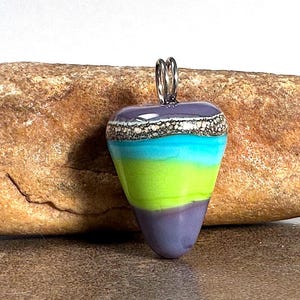 May include: A teardrop-shaped glass pendant featuring horizontal bands of purple, lime green, and bright turquoise, topped with a textured white and black speckled accent and a silver-toned metal bail for hanging.