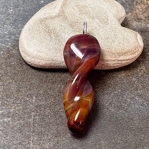 May include: A twisted, teardrop-shaped pendant in shades of red, amber, and purple. The glass pendant has a silver bail and is resting on a light-colored stone against a textured gray background. The pendant is a unique piece of jewelry.