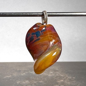 May include: A teardrop-shaped glass pendant featuring a swirling blend of amber, orange, and deep purple colors with dark blue accents. The pendant has a smooth, glossy finish and is suspended from a silver-toned metal jump ring on a thin rod.