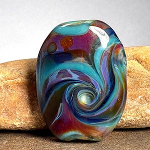 May include: A smooth, oval-shaped glass bead featuring a swirling pattern of deep purple, vibrant turquoise, and amber tones. The surface has a glossy finish with a central vortex design that blends the colors together in a fluid, circular motion.