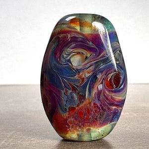 May include: A colorful, oval-shaped glass bead with swirling patterns of blue, purple, red, and yellow. The bead has a glossy finish and a unique, artistic design, suitable for jewelry making or decorative purposes.
