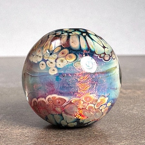 May include: A spherical glass paperweight featuring a swirling pattern of blue, purple, and orange hues. The surface displays intricate, cell-like organic shapes in cream and brown tones, with a reflective finish and a bright white light reflection.