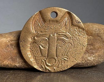 May include: A round, bronze-colored pendant featuring a detailed relief of a wolf's face. The pendant has a hole at the top for hanging and is resting on a light brown stone. The wolf's features are intricately carved.