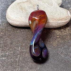 May include: A handcrafted glass pendant with a swirling design in shades of red, orange, purple, and blue. The pendant has a twisted shape and a silver-colored bail for attaching to a chain. The bottom of the pendant features dark purple and brown circles.