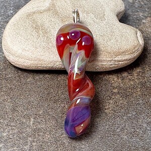 May include: A twisted glass pendant with a silver bail. The pendant features a red top with three purple dots, transitioning to a red and purple swirl, and ending in a purple base. The pendant is approximately 3 inches long.