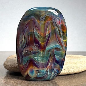 May include: A colorful, oval-shaped glass art piece with swirling patterns in shades of blue, purple, orange, and yellow. The glass has a glossy finish and appears to be resting on a light-colored stone. The piece has a unique, artistic design.