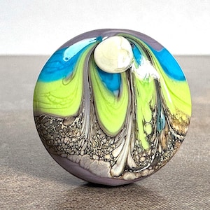 May include: A circular glass bead featuring a swirling pattern of bright lime green, vibrant turquoise, and muted gray tones. The bottom section displays a textured, pebbled appearance in shades of brown and tan, topped with a small, smooth white orb.