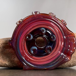 May include: A handcrafted glass art piece with a spiral design. The piece features a blend of red, purple, and blue hues, with small, round, blue accents. The glass is set on a natural stone, showcasing its unique shape and color variations.
