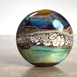 May include: A spherical glass paperweight featuring a layered design with a smooth, glossy finish. The surface displays horizontal bands of deep brown, iridescent blue, a textured white and black cell pattern, and a translucent mint green.