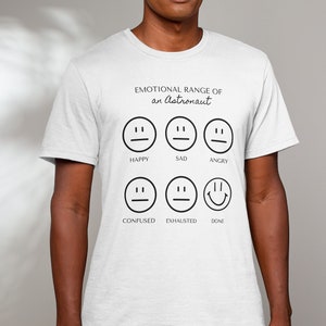 May include: White t-shirt with the text "EMOTIONAL RANGE of an Astronaut" above a series of emoji faces. The faces represent emotions: happy, sad, angry, confused, exhausted, and done. The shirt is worn by a person.