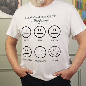 Puede incluir: Camiseta blanca con el texto "EMOTIONAL RANGE OF a Professor" y seis caras emoji que representan diferentes emociones. La persona que lleva la camiseta tiene barba blanca y está de pie frente a obras de arte enmarcadas.