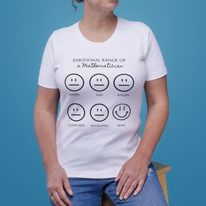Puede incluir: Una camiseta blanca con el texto "EMOTIONAL RANGE of a Mathematician" y varias caras sonrientes que representan diferentes emociones. La persona lleva vaqueros azules y está sentada en un taburete de madera sobre un fondo azul.