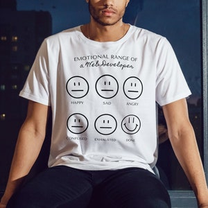 May include: White t-shirt with the text "EMOTIONAL RANGE OF a Web Developer" and six different facial expressions representing emotions. The expressions include happy, sad, angry, confused, exhausted, and done. The person in the image is wearing black pants.