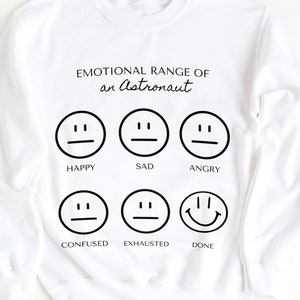 May include: White sweatshirt with the text "EMOTIONAL RANGE OF an Astronaut" above six different emoji faces representing emotions: happy, sad, angry, confused, exhausted, and done. The sweatshirt is a crew neck style.