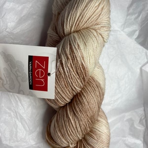 May include: A skein of Zen Yarn Garden yarn in a natural color palette of cream and brown. The yarn is tightly wound and has a subtle gradient effect. A tag with the Zen Yarn Garden logo is attached.
