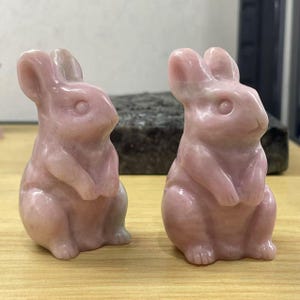 May include: Two pink rabbit sculptures carved from polished pink opal. The rabbits are sitting upright with their paws in front of them.