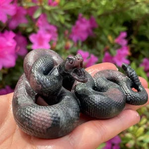 May include: A black and pink carved stone sculpture of a rattlesnake with its mouth open. The sculpture is detailed and realistic.
