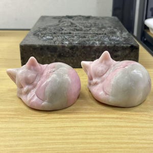 May include: Two small, sleeping cat sculptures carved from pink and grey stone. The cats are curled up in a sleeping position with their eyes closed.