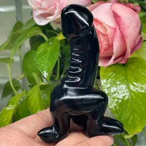Könnte beinhalten: Eine schwarze Obsidian-Skulptur einer hockenden Figur mit einem runden Kopf und einem erhobenen Arm. Die Skulptur ist poliert und hat eine glatte, glänzende Oberfläche.