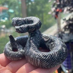May include: A detailed, coiled snake figurine in shades of grey and white. The sculpture has a textured, scaled appearance, with intricate patterns. The snake is posed in a striking, upright position, showcasing its detailed features. The figurine is held in a hand.