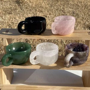 May include: Five small cups made of different types of stone. The cups are in a row on a wooden shelf. The cups are black, pink, green, white, and purple.