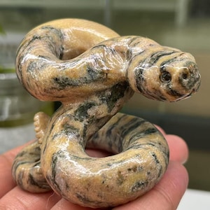 Op de afbeelding: Een handgesneden stenen sculptuur van een slang, met een gemarmerd patroon in lichtbruin, roze en zwart. De slang is opgerold in een verdedigende positie, met zijn kop omhoog en zijn ogen dicht.