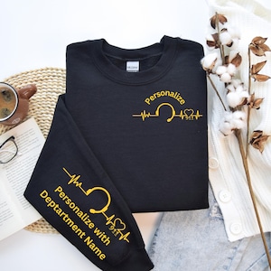 May include: Black sweatshirt with gold text and a design featuring a heartbeat, a headset, and the number 911. The text reads "Personalize" and "Personalize with Department Name" on the sleeve. The sweatshirt is a Gildan brand.
