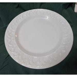 May include: A round white ceramic dinner plate featuring a smooth, flat centre and a wide, raised rim decorated with an embossed, repeating basket-weave pattern that encircles the entire edge of the plate, creating a textured border.