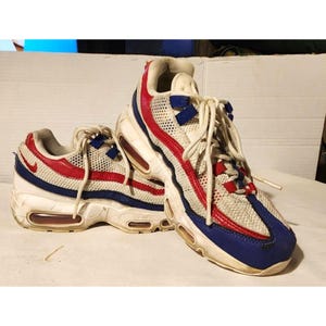 Puede incluir: Par de zapatillas deportivas blancas, rojas y azules. Las zapatillas presentan una parte superior de malla con detalles rojos y azules, una entresuela blanca y una unidad de aire visible. Cordones blancos.