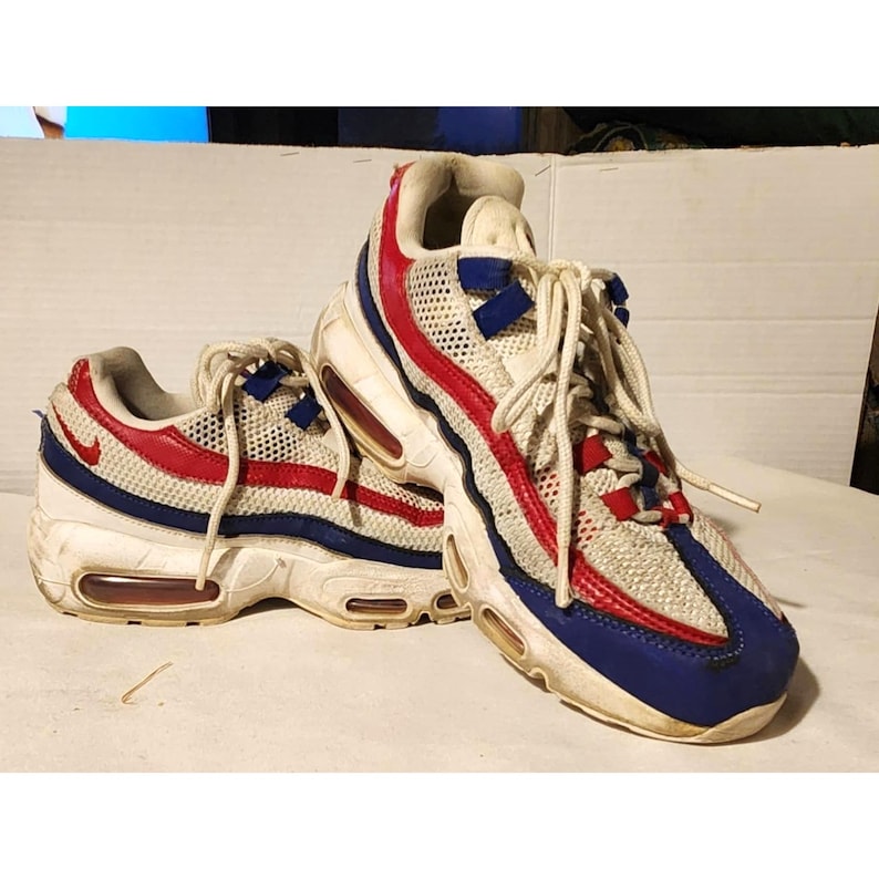 Puede incluir: Par de zapatillas deportivas blancas, rojas y azules. Las zapatillas presentan una parte superior de malla con detalles rojos y azules, una entresuela blanca y una unidad de aire visible. Cordones blancos.