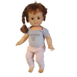 Puede incluir: Muñeca vintage con cabello castaño peinado en una trenza lateral. La muñeca viste un body gris con el texto "Happiest little one" y pantalones rosas. La muñeca está de pie con los brazos a los lados.
