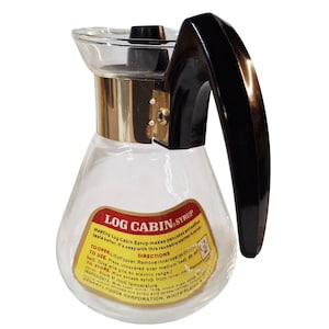 May include: A clear glass syrup warmer with a black plastic handle and a metallic collar. The label reads LOG CABIN SYRUP with directions for heating, storage, and ingredients. The container features a wide base and a narrow top opening.