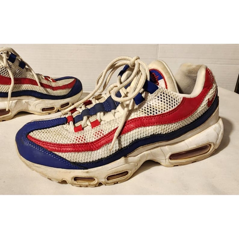 Puede incluir: Par de zapatillas deportivas blancas, rojas y azules. Las zapatillas presentan una parte superior de malla con detalles en rojo y azul, una entresuela blanca y amortiguaci&oacute;n de aire visible. Los cordones son blancos.