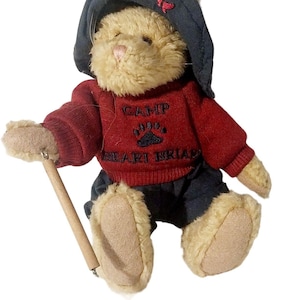 May include: A plush brown bunny wearing a blue hat with a red butterfly, a red sweater with the text "CAMP BEAR BRIAR" and blue pants. The bunny is holding a wooden stick.