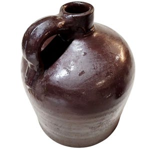 May include: A dark brown glazed ceramic jug with a rounded body and a single side handle. The vessel features a smooth, glossy finish on the upper portion and a slightly lighter, matte-textured base, with a narrow neck and a circular opening.