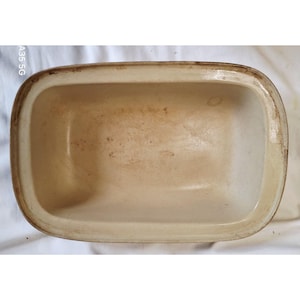 May include: A rectangular, off-white ceramic baking dish with rounded corners and a slightly raised rim. The dish has a light brown rim and shows signs of wear and use, with some discoloration on the interior surface. The dish is empty.