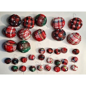 May include: A collection of Christmas ornaments in various sizes. The ornaments are round and covered in festive fabrics, including plaid patterns and snowflake designs. Colors include red, green, and white. Each ornament has a small wire loop for hanging.
