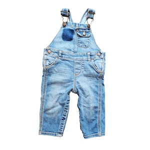 May include: Light blue denim overalls for a baby or toddler. The overalls have adjustable straps, a bib pocket, and two front pockets. A dark blue patch is sewn on the bib. The overalls are made of a light-wash denim material.