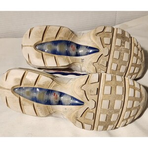 Puede incluir: Par de zapatillas deportivas blancas y azules, vistas desde las suelas. Las suelas muestran un patr&oacute;n texturizado y desgastado con detalles azules y rojos. Las zapatillas parecen usadas.